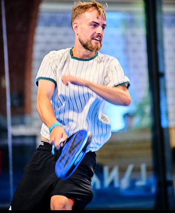 Junger Mann mit weiß/grünem T-Shirt und schwarzer Hose in Nahaufnahme beim Padel spielen