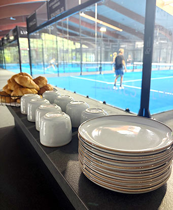 Im Vordergrund stehen Teller und Tassen neben ein paar Croissants. Im Hintergrund ein Padel-Court