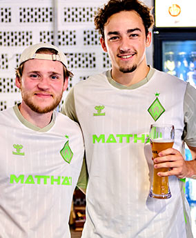 Zwei Männer im Werder-Trikot stehen nebeneinander. Der rechte hält ein Weizenbier in der Hand